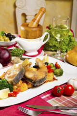 Two fried carp steaks served on a white plate with boiled potatoes and broccoli next to fresh vegetables, sauce and glass of white wine.