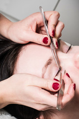 Hands holding forceps over young beautiful girl is getting eyelash extensions closeup. 