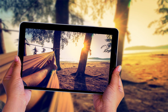 Human hand take picture with tablet of feet sleeping on swing at the sunset beach background.