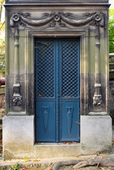 Naklejka premium Rectangular blue double door made of steel. Ornaments of vases, floral and cross pattern. The ornate door frame contains chiseled relief of torches carved in stone. An old tomb / crypt at a graveyard.