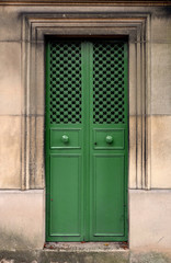 Green closed iron double door with door knobs and a hole pattern in a stone wall. The door frame is rectangle, ornated by chiseled lines. A texture of small crosses which are connected by steel bars.