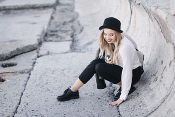 blonde in a black pants and a hat and white shirt sitting on the floor and smiling