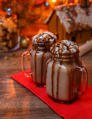 Two glasses of cocoa with whipped cream and chocolate syrup on wooden table and Gingerbread house, cookie jar and christmas tree with toys and garland on background.