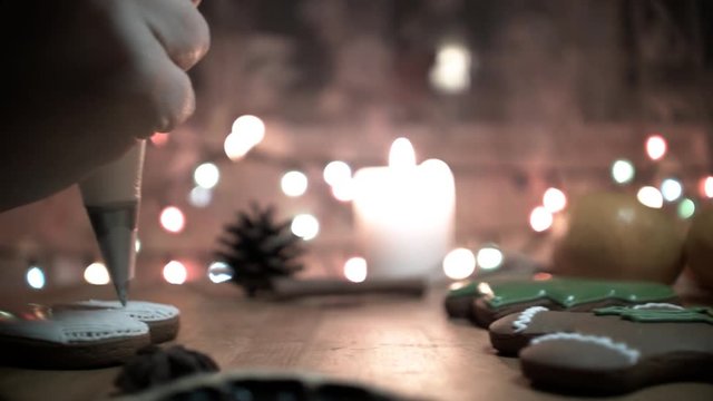 Female hand decorate Gingerbread white heart cookie with food icing on the table, close up. Candle, pine cone, mandarin, canella, bokeh in backgound.