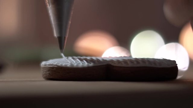 Female hand decorate Gingerbread white heart cookie with food icing on the table, close up. Candle, pine cone, mandarin, canella, bokeh in backgound.