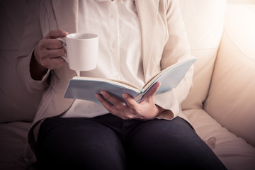 woman is sitting on a sofa and reading a book while holding a co