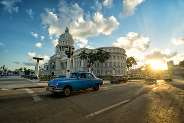 Blue retro car is riding near ancient colonial Capitol building at the center of Havana at sunset 