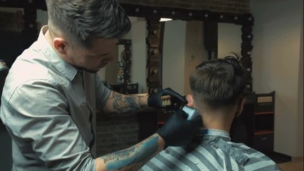 Attractive barber with dark hair wearing white shirt, watch and black gloves doing a haircut for client with scissors at barber shop,