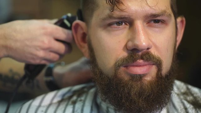 Creating new hair look. Young bearded man getting haircut by hairdresser while sitting in chair at barbershop