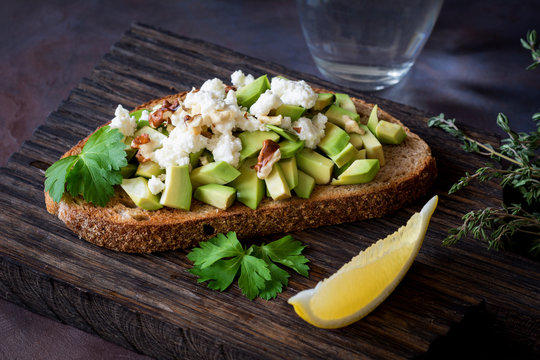 Avocado, cheese and walnuts salad on toasted bread, served on wooden cutting board with lemon wedge. Healthy snack