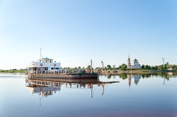 Ferryboat with people and cars goes on Vychegda River with ancient cathedral on riverside in background. Solvychegodsk, Arkhangelsky region, Russia.