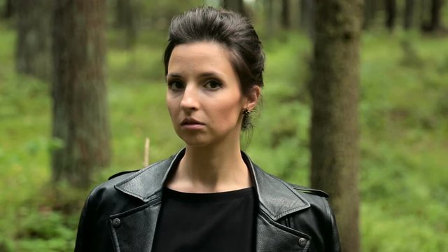 young brunette woman with makeup, in a leather black jacket and dress sitting on a tree in a pine forest in summer