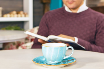 Young beautiful man sitting and drinks coffee. Also he reading a book