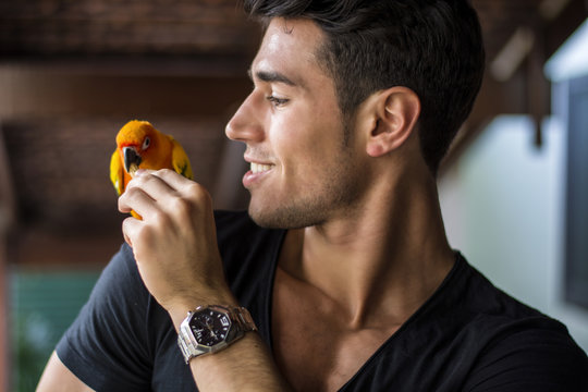 Smiling handsome man feeding parrot sitting on his shoulder. Horizontal indoors shot