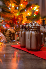 Two glasses of cocoa with whipped cream and chocolate syrup on wooden table and Gingerbread house, cookie jar and christmas tree with toys and garland on background.