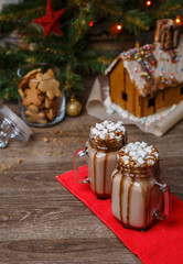 Two glasses of cocoa with marshmallows, whipped cream and chocolate syrup on wooden table and Gingerbread house, cookie jar and christmas tree with toys and garland on background.