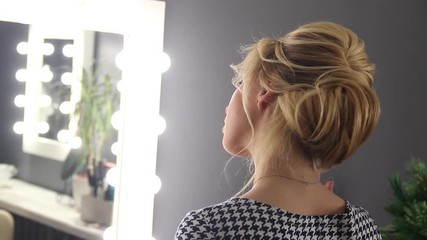 Attractive blonde girl looks at the mirror with bulb lamps in the beauty studio. Concept of a satisfied customer of make-up and hairstyle