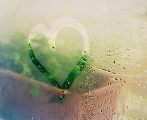 Dibujo de un corazón en el vapor de una ventana con una maceta  con plantas de fondo 