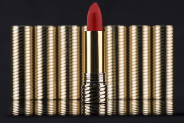 Dark red color lipstick sample in a sharp golden tube, with other blurred golden tubes on a black...