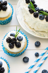 Homemade cupcakes and cake with frosting, blueberries, blackberries and rosemary leaves on white wooden background