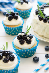 Homemade cupcakes and cake with frosting, blueberries, blackberries and rosemary leaves on white wooden background