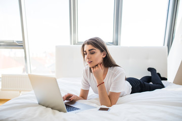 Portrait of beautiful happy young woman lying in bed and using laptop at home