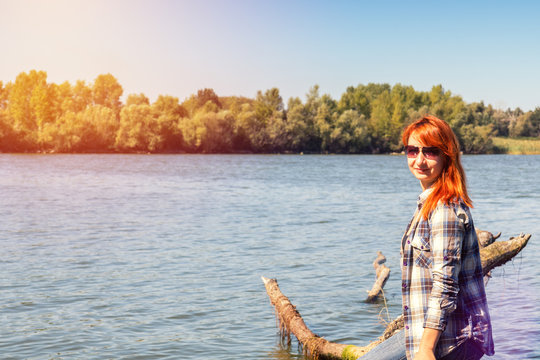 Young woman with red hair in checkered shirt and sunglasses at the river in the sunny day. Coloring and processing photo with soft focus in instagram style.