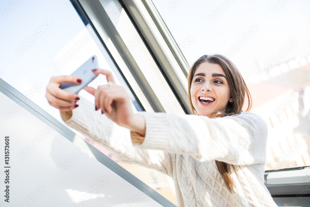 Wall mural young beautiful caucasian woman near window, making faces, smiling and making selfie the morning.