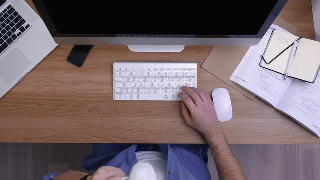 Overhead high angle shot of Caucasian worker in casual clothes drinking coffee while working at computer In modern office. 4K UHD