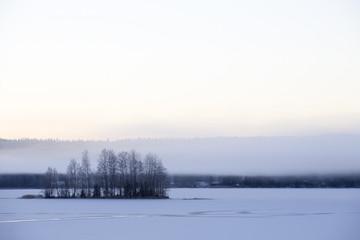 Foggy winter morning. Small island. Ice covered with snow. Foggy mountains in the background.