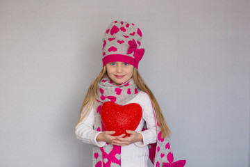 cute little girl in a cap and a scarf with a heart holding a red heart. Valentine's day holiday.