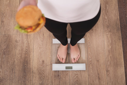 Diet And Fast Food Concept. Overweight Woman Standing On Weighing Scale Holding Burger ( Hamburger ). Unhealthy Junk Food. Dieting, Lifestyle. Weight Loss. Obesity. Top View