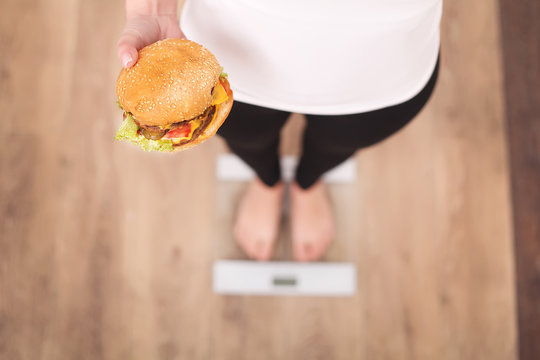Diet And Fast Food Concept. Overweight Woman Standing On Weighing Scale Holding Burger ( Hamburger ). Unhealthy Junk Food. Dieting, Lifestyle. Weight Loss. Obesity. Top View