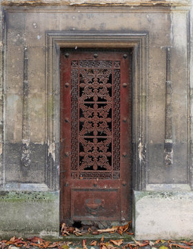 Old and weathered red / brown closed iron door in a stone wall of an old tomb / crypt at a graveyard. Artistic curvy flower ornaments and geometric forms, surrounded by a rectangle border line frame.