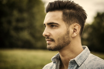 Handsome young man at countryside, in front of field or grassland, wearing shirt, looking away to a side