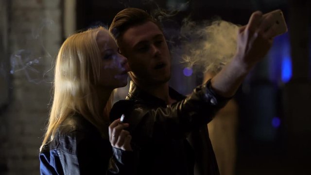 Young man and woman smoking and making selfie on smartphone at nightclub