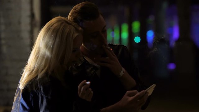 Young couple smoking cigarettes and looking through photos on smartphone