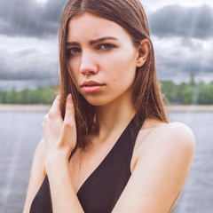 Model - cute brunette with green eyes, wearing a dress. Girl standing in the rain on a background of the river / trees. The sky is covered with clouds / storm / storm cloud. He looks into the camera.