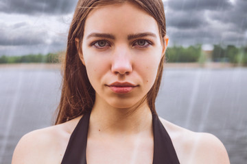 Model - cute brunette with green eyes, wearing a dress. Girl standing in the rain on a background of the river / trees. The sky is covered with clouds / storm / storm cloud. He looks into the camera.