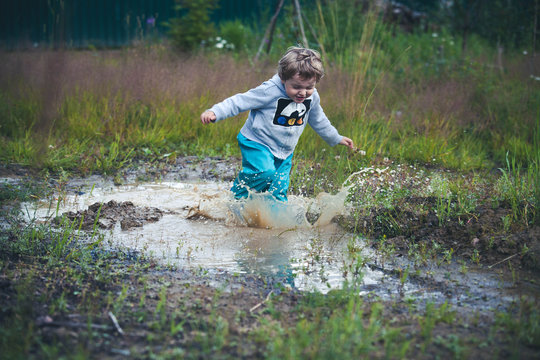 Cute curly baby dressed in boots and rubber pants jumping in a puddle. Funny kid with children's happiness.