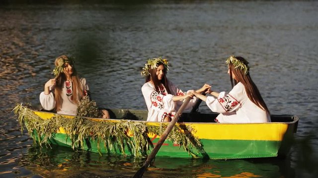 Three beautiful girls in the Slavic clothes in a boat floating on the river. Two girls rowing oars on the water. girl with long hair