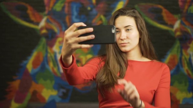 Young beautiful girl in a red dress makes selfie on your smartphone. In room. Slow motion.