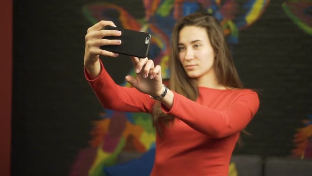 Young beautiful girl in a red dress makes selfie on your smartphone. In room. Slow motion.