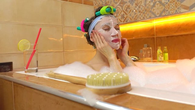 Brunette woman applying face mask in foamy bath. Beauty treatments at home. 4K shot