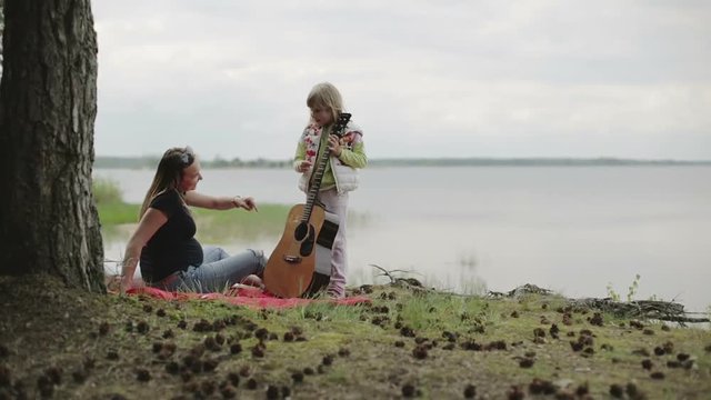 Daughter plays the guitar for his mother on the shore of the lake in cloudy weather.