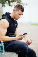 Fitness man wearing sportswear listening to music sitting on a stone bench. Relax after  hard workout, talking.
