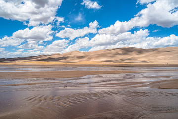 White spring clouds, on blue sky, passing over rolling hills of sand dunes and gently flowing Medano Creek. Great Sand Dunes National Park & Preserve, Colorado, USA.