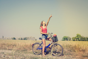 Beautiful pinup girl having fun cycling in fields under bright blue summer sky and pointing up on...