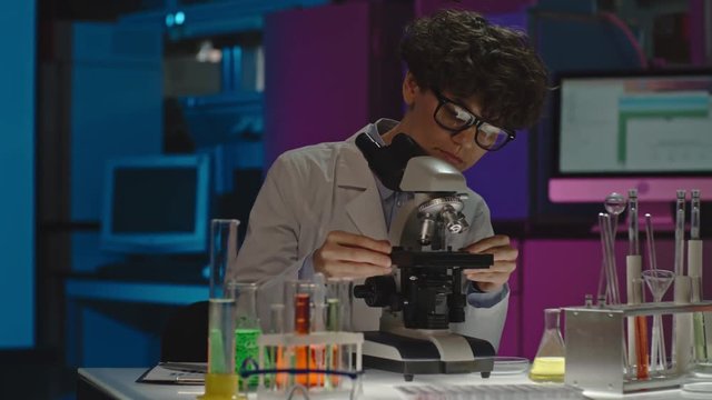 PAN of curly female scientist in glasses making notes on clipboard and looking into microscope in laboratory, then checking something on tablet 