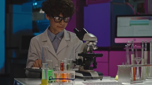 PAN of concentrated curly female scientist in glasses making notes on clipboard and studying sample in microscope, then checking something on tablet 